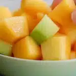 Hand picking a cube of mango from a fresh mango melon fruit salad in a bowl.