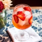 Strawberry spritz cocktail in a stemmed glass with ice and fresh sliced strawberries on a floral summer table